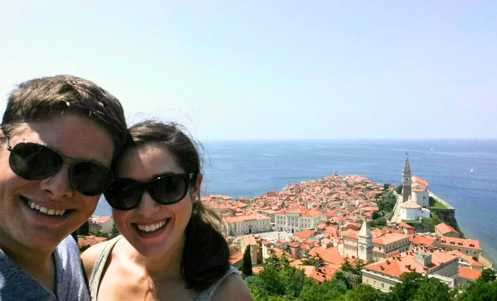 kate storm and jeremy storm taking a selfie from the piran town walls, with cityscape of piran slovenia and adriatic sea visible in the background