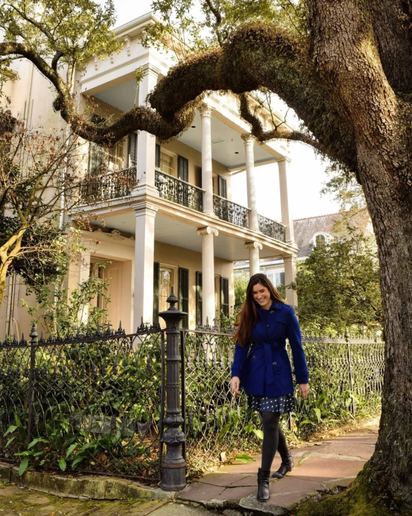 Kate Storm in a blue coat walking in front of a mansion and below a large oak tree in the Garden District during 3 days in NOLA new orleans