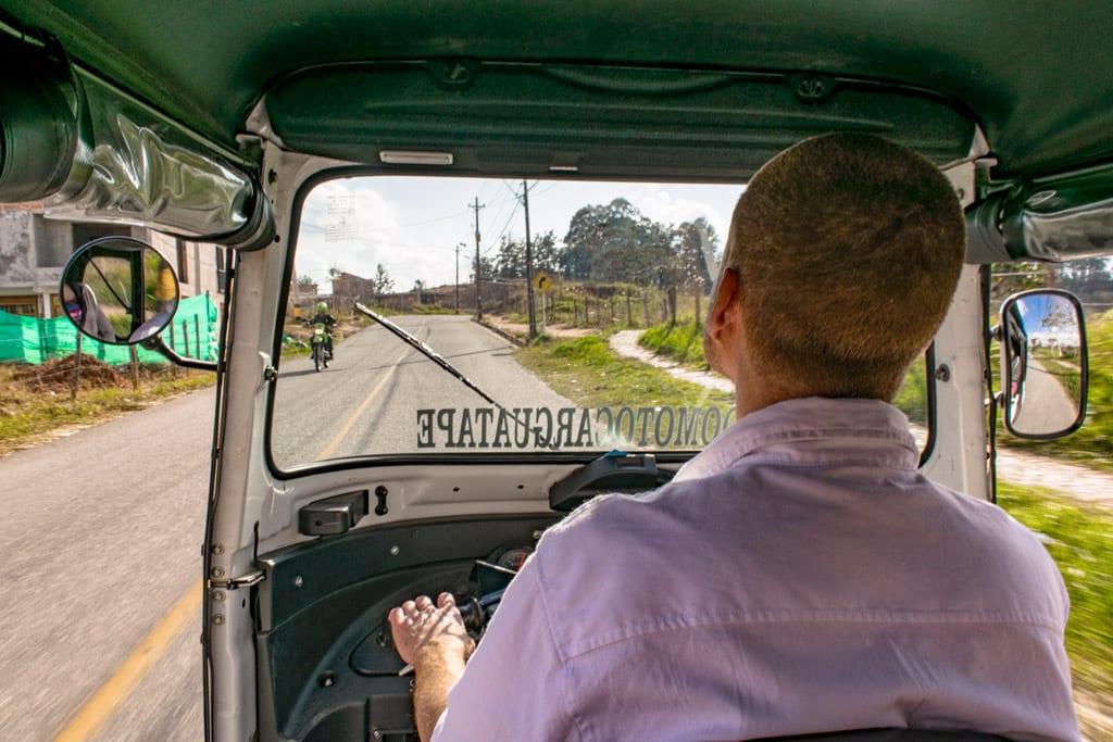 view of a tuk tuk and driver traveling down the road in guatape colombia, as seen from the back of the tuk tuk