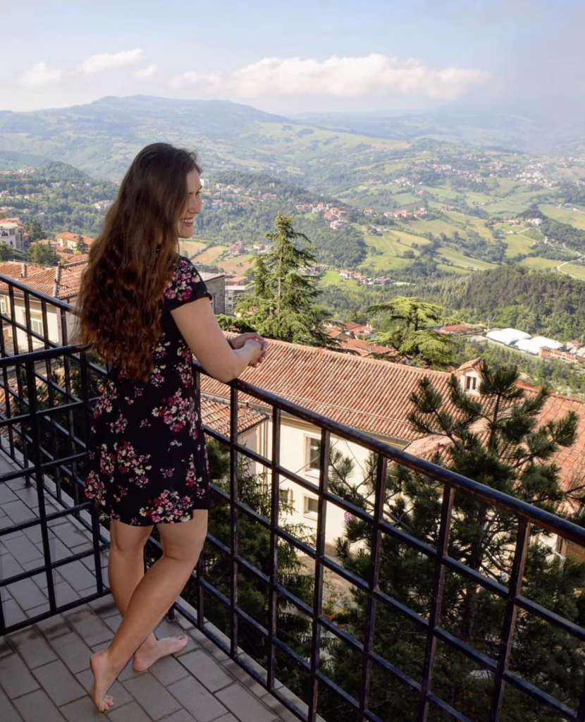 kate storm in a black floral dress standing on a balcony at hotel la rocca in san marino with countryside visible behind her