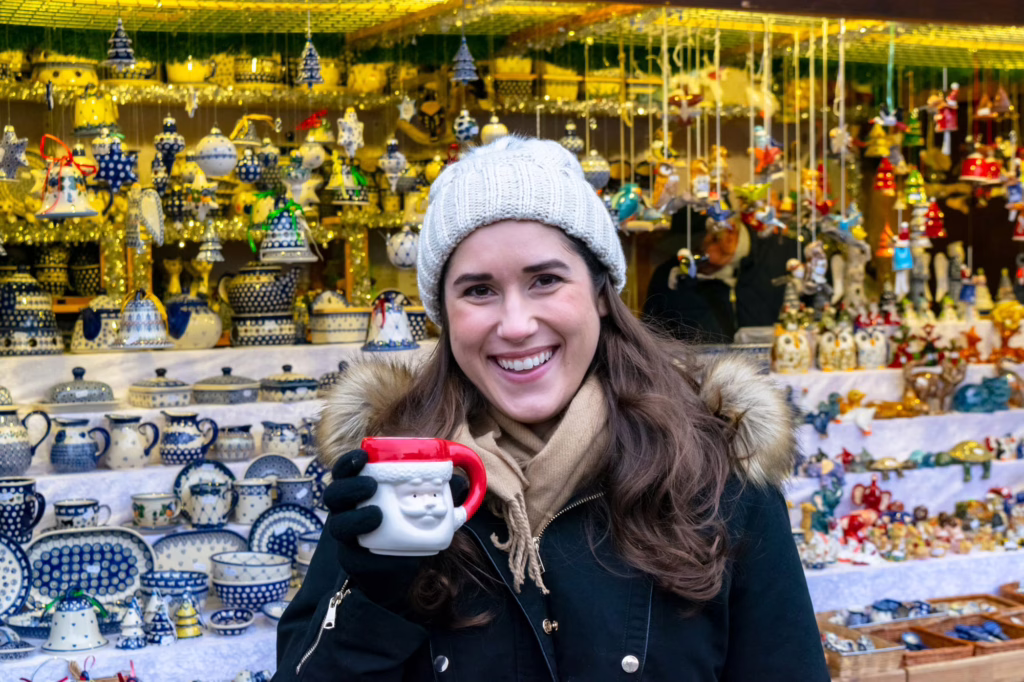 Kate Storm at a Christmas market in Vienna wearing a cream knit hat and holding up a Santa mug