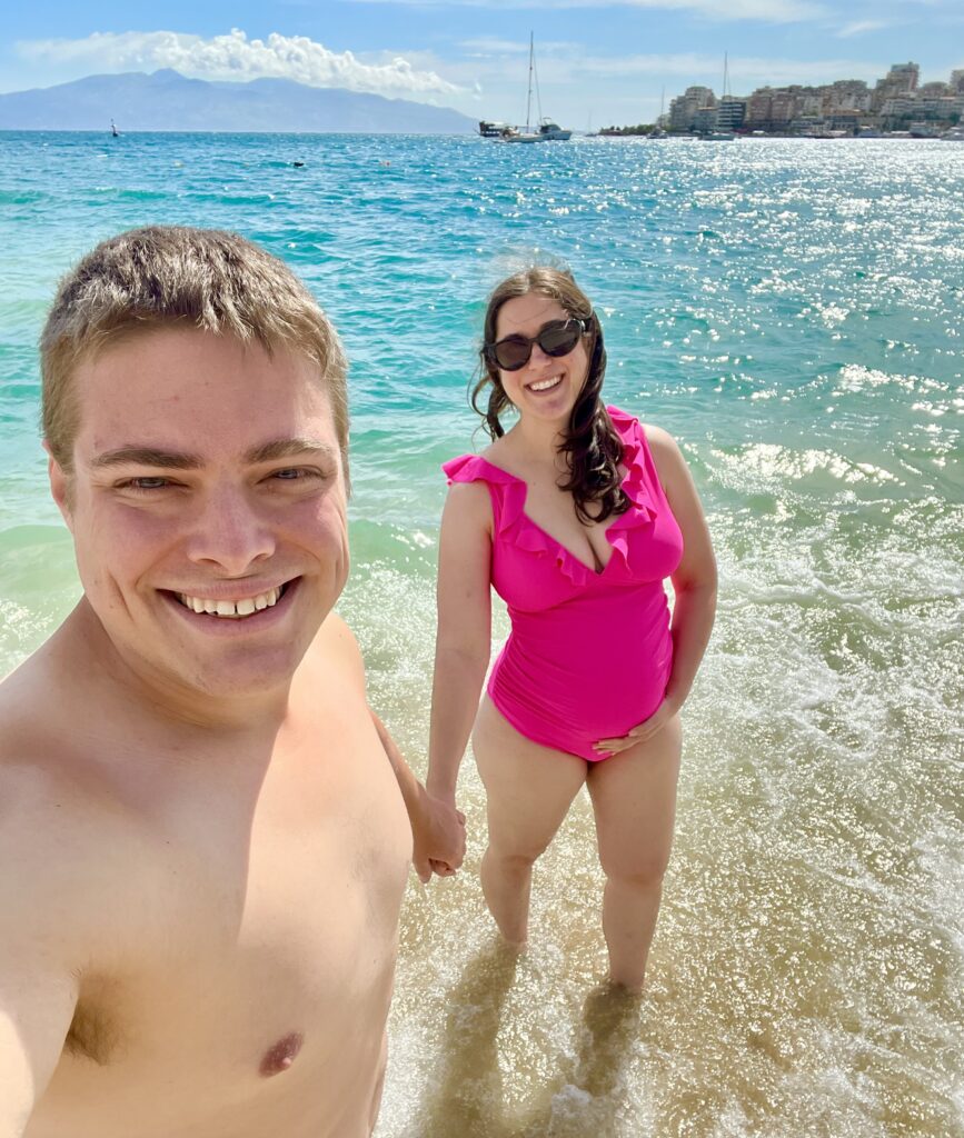 kate storm and jeremy storm standing in shallow water on the beach in saranda albania, taking a selfie and wearing bathing suits