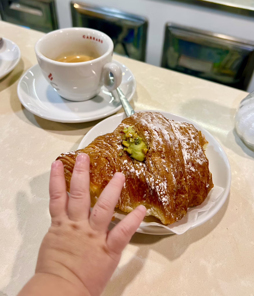 young baby's hand reaching out to a pistachio cornetto on a coffee bar in italy with caffe visible behind it