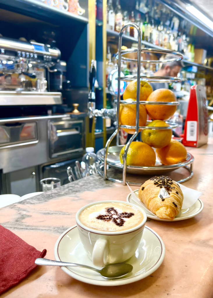 italian coffee sitting on a bar with a pastry, group of oranges, and espresso machine visible behind it