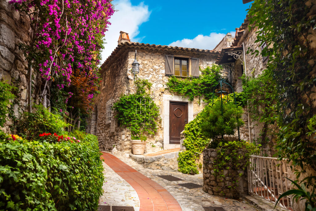 small street in eze france lined with stone buildings and purple flowers blooming on one side of the street