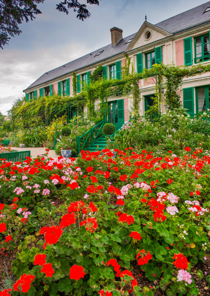 pink facade of monet house in giverny france on a cloudy day with red and pink flowers blooming in the foreground
