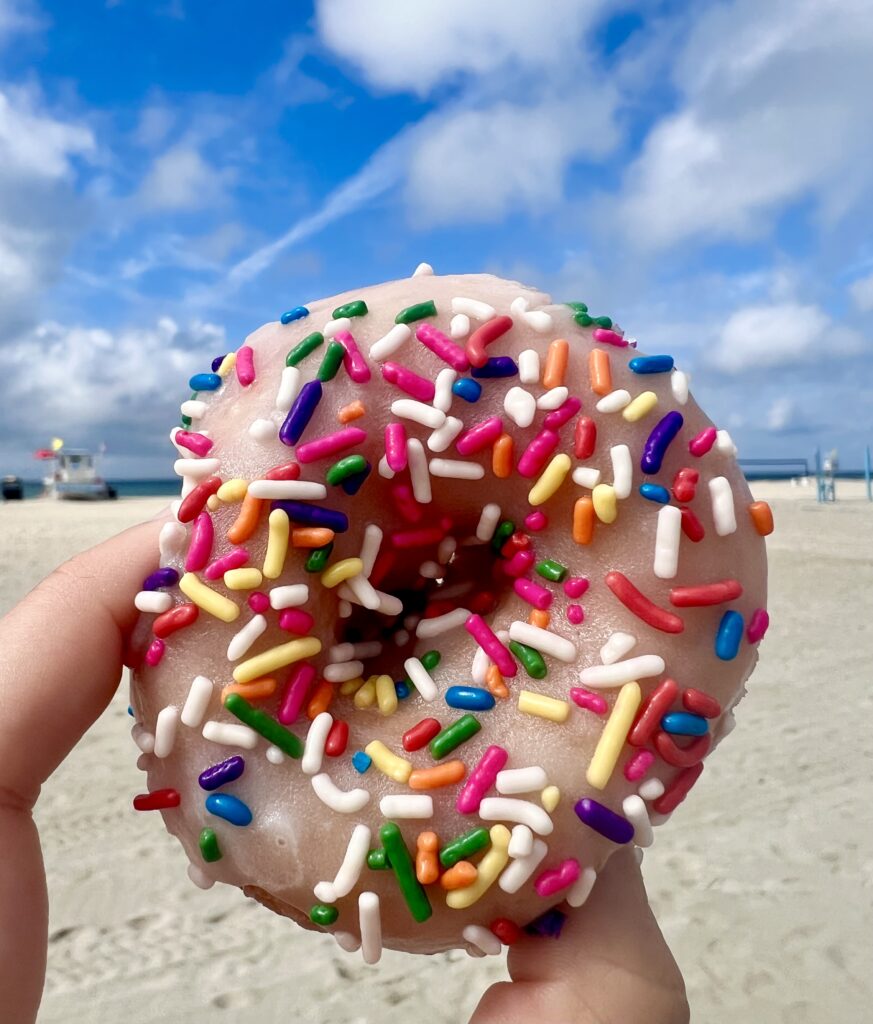 sprinkle donut with vanilla frosting from hang 10 donuts atlantic beach nc being held up in front of sandy beach