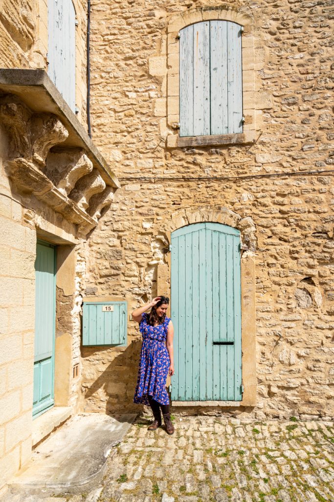 kate storm in a blue dress posing in front of a stone building with light blue shutters and doors in gordes france, one of the best places in the luberon valley to visit