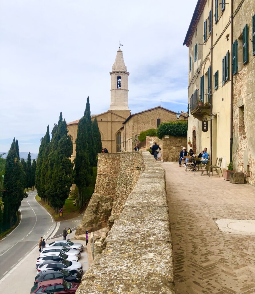 view of pienza italy walls with church steeple in the background, a road with cars parked below to the left, and a stone walkway on the right