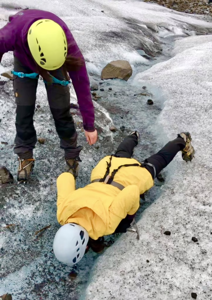 kate storm in a yellow raincoat balancing over an ice stream to drink glacier water in iceland, with a guide hovering over her