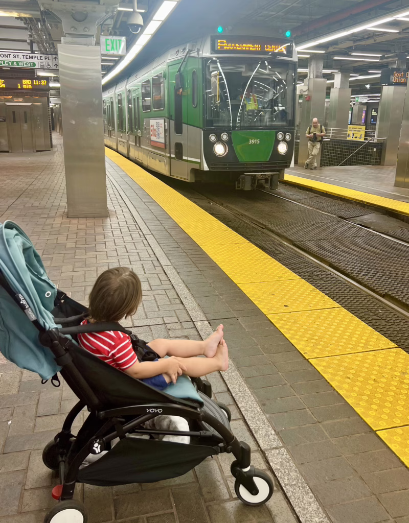 toddler sitting in a stroller looking at the green line of the t in boston ma as it approaches the station