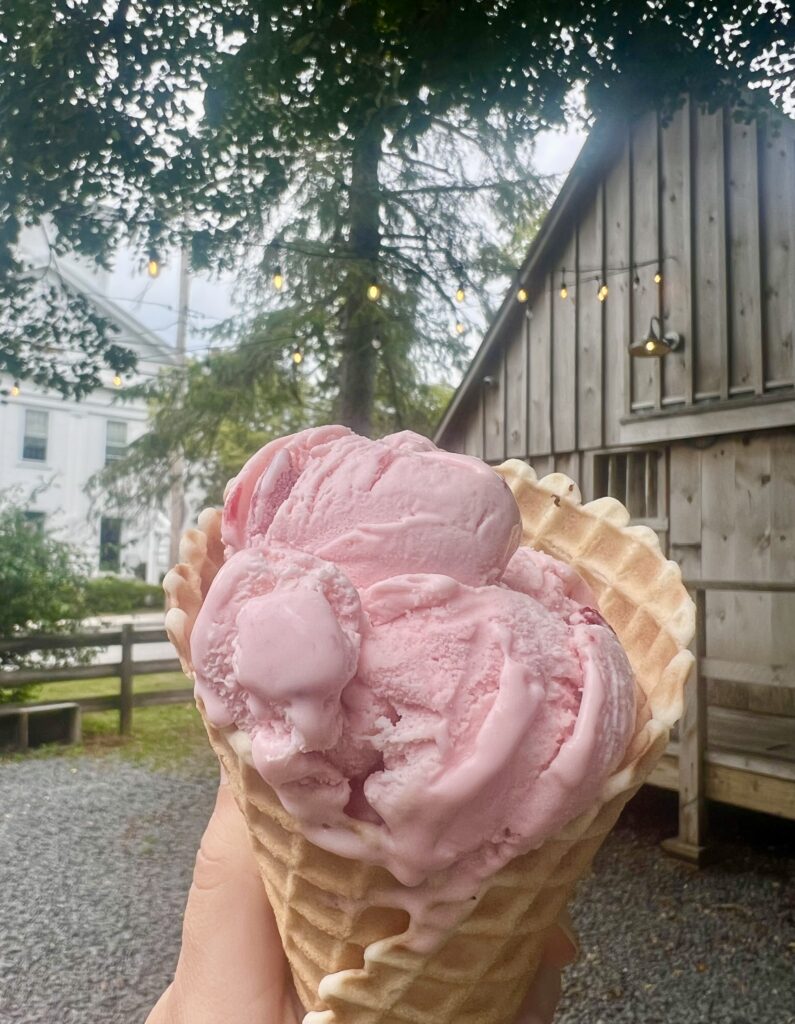 strawberry ice cream cone being held up in front of fairy lights, an aged barn and a white new england church at bramhalls country store plymouth ma