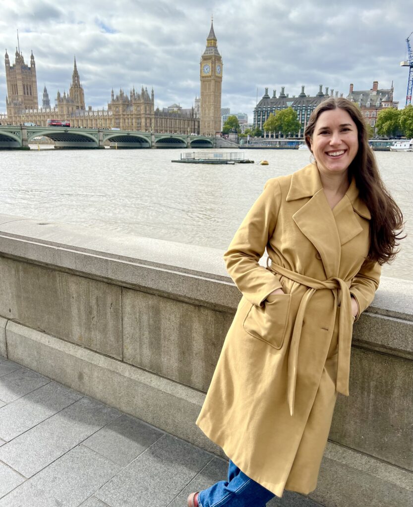 kate storm in a tan coat standing in front of elizabeth tower house of parliament and thames in london england