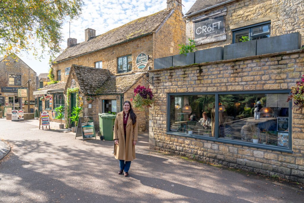 kate storm standing in front of a collection of stone buildings in the center of bourton on the water cotswolds village