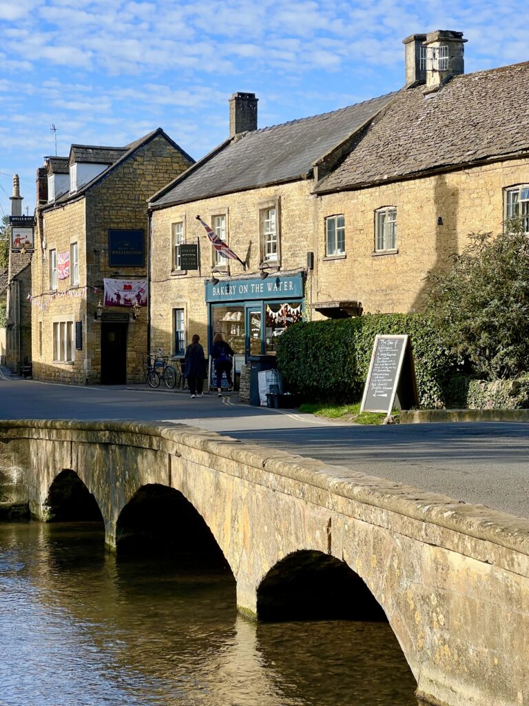 front facade of bakery on the water visible across the river windrush and a stone bridge in the small cotswold village bourton on the water
