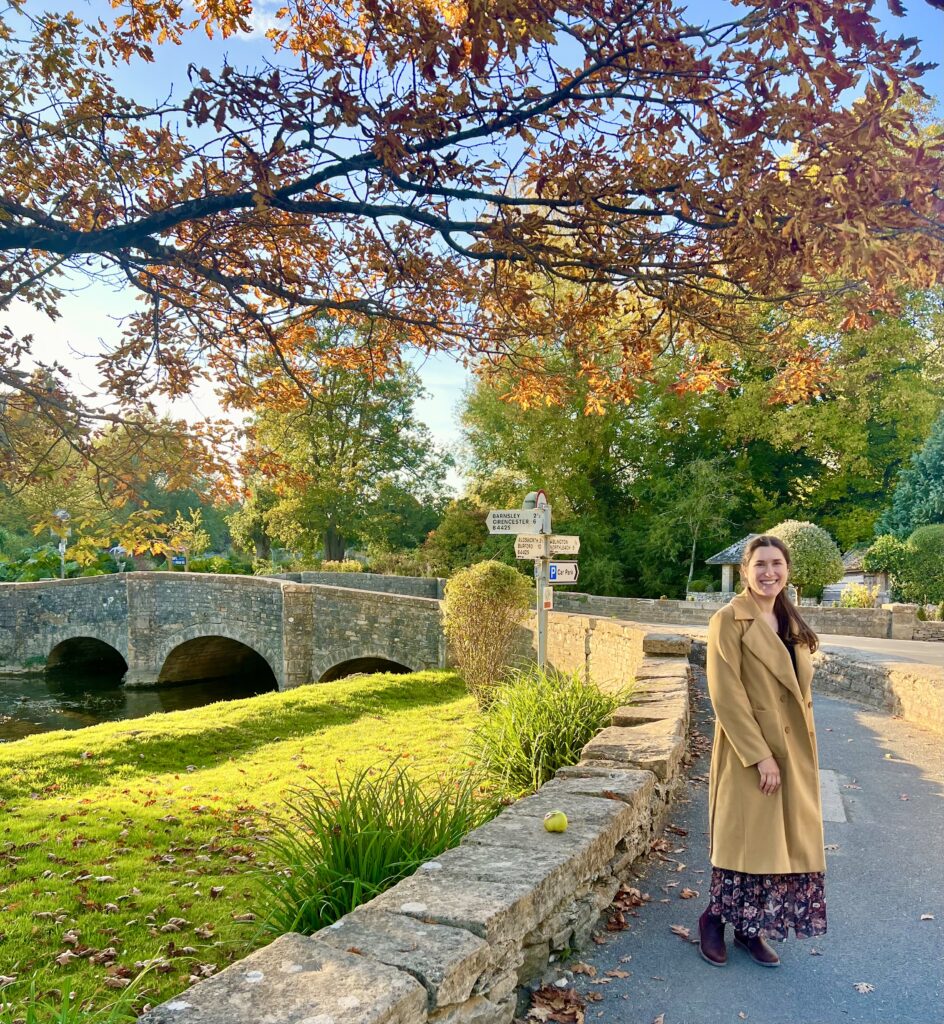 kate storm standing in bibury in front of a stone bridge and under a tree with orange foliage in october