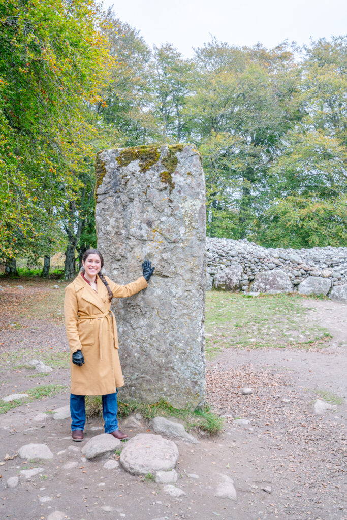 kate storm in a brown coat standing with her hand touching a standing stone at the clava cairns, one of the best places to visit near inverness scotland