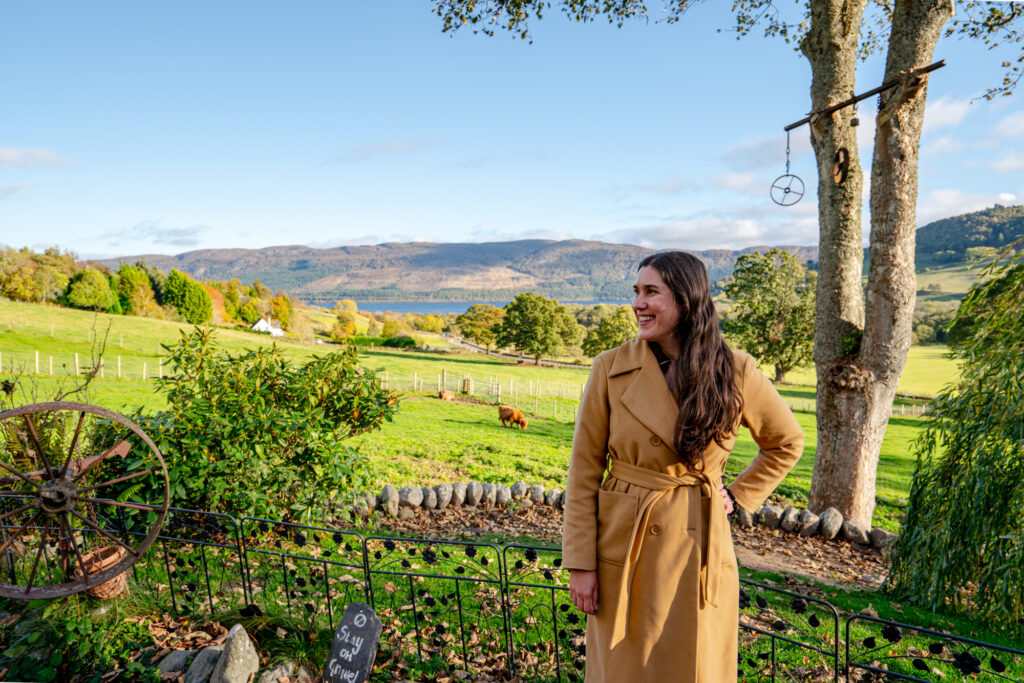 kate storm in a tan coat standing on a farm in scotland with hairy coos behind her