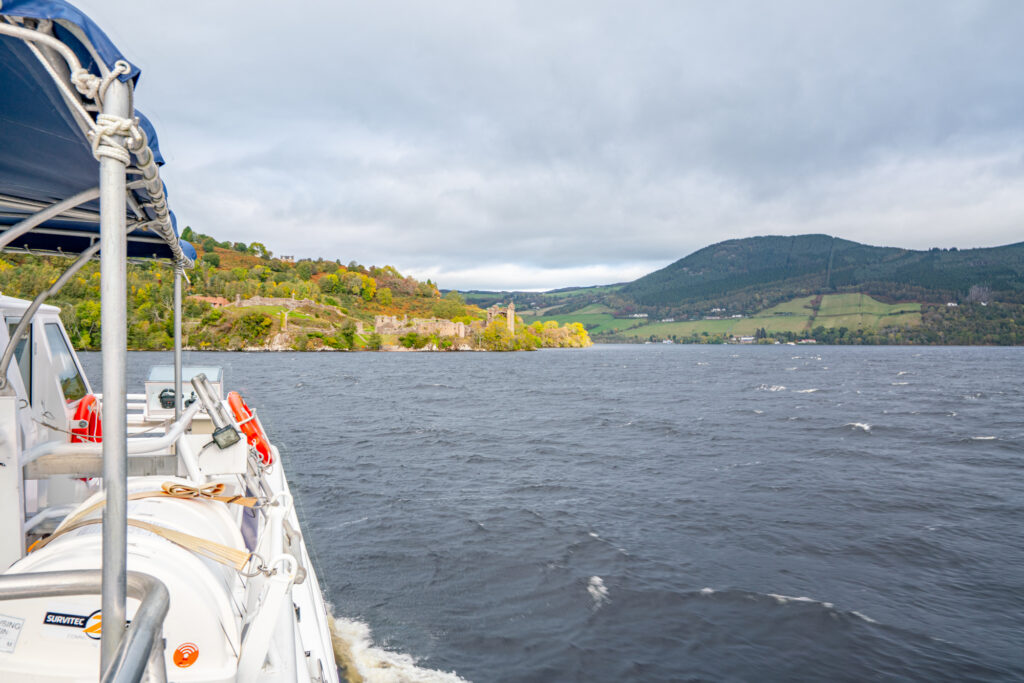 view of the side of a white boat on loch ness with urquhart castle visible in the distance beyond the lake