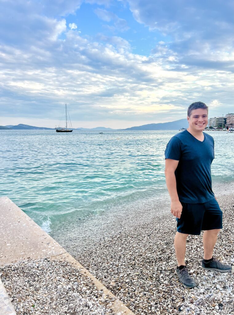 jeremy storm standing on the beach in saranda albania near the shore with sailboat visible behind him