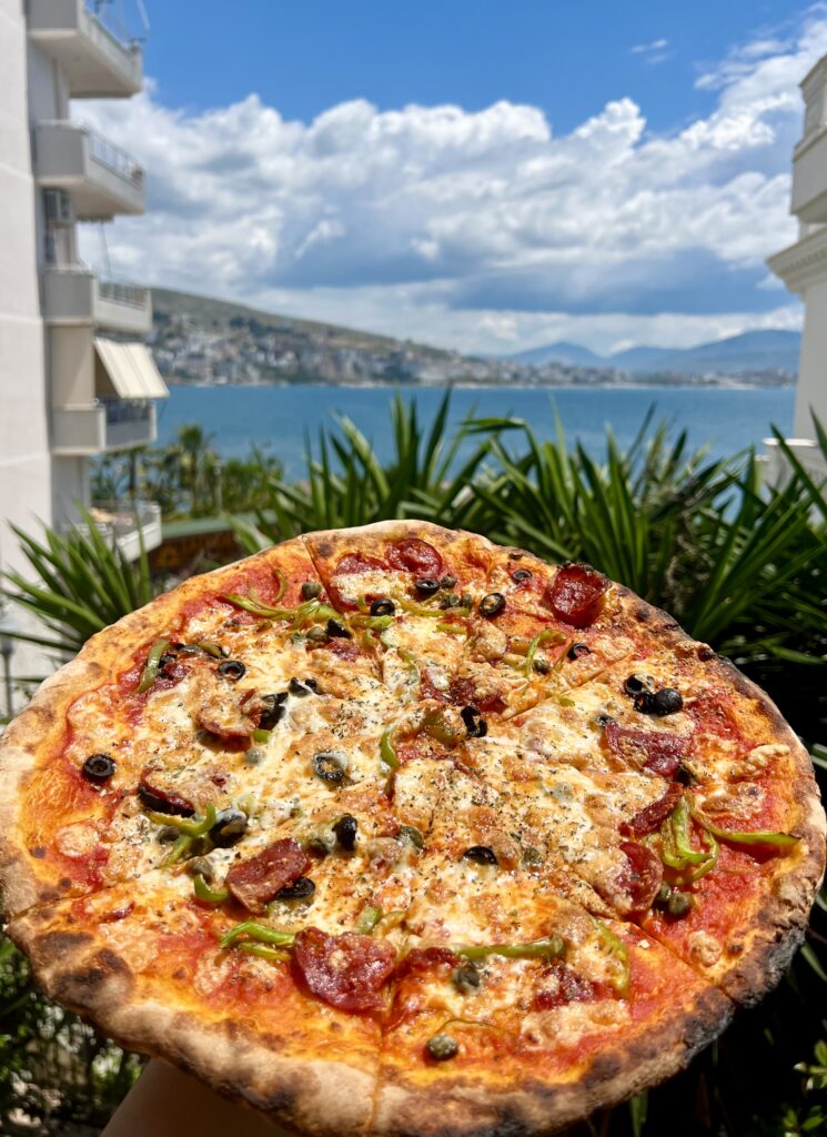 view of a pizza being held up to a view of the sea in saranda albania