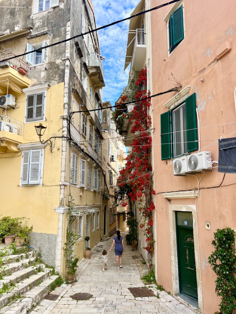 kate storm and her young toddler walking away from the camera and down a narrow, colorful street in corfu greece
