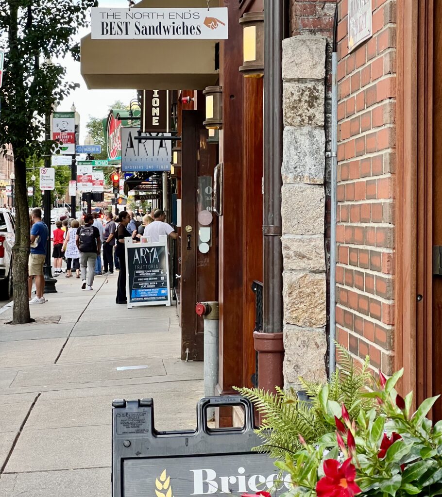 view of sidewalk in boston little italy lined with people and signs advertising restaurants and bakeries
