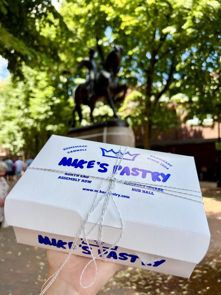 white and blue pastry box from mike's pastry being held up at paul revere mall in front of statue