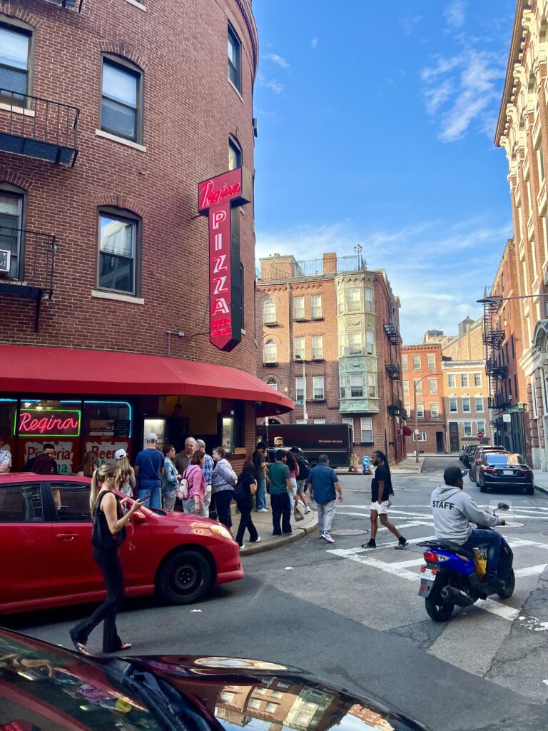 intersection in the boston north end with person riding a scooter in the foreground and the facade of regina pizzeria in the background, one of the legendary north end restaurants