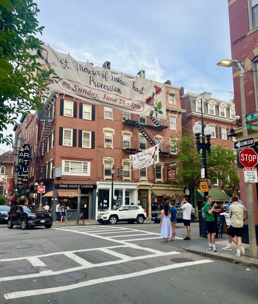 view of hanover street north end being approached from the corner, a banner hanging across the street is advertising a north end summer feast, one of the best things to do in the north end boston