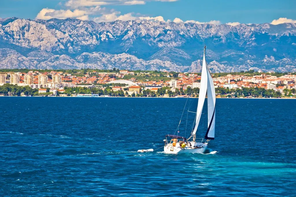sailboat on the water with city of zadar croatia in the background and mountains behind it