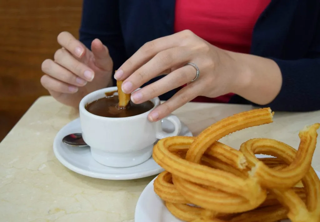 Kate Storm's hands shown dipping a churro into chocolate with a stack of churros off to the side--even with only a few days in Madrid, eating lots of churros is a msut!