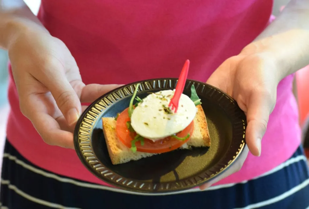 Kate Storm in a pink shirt holding a plate with a small tapa on it made of bread, tomato, and cheese. Photo is focused on the tapa.