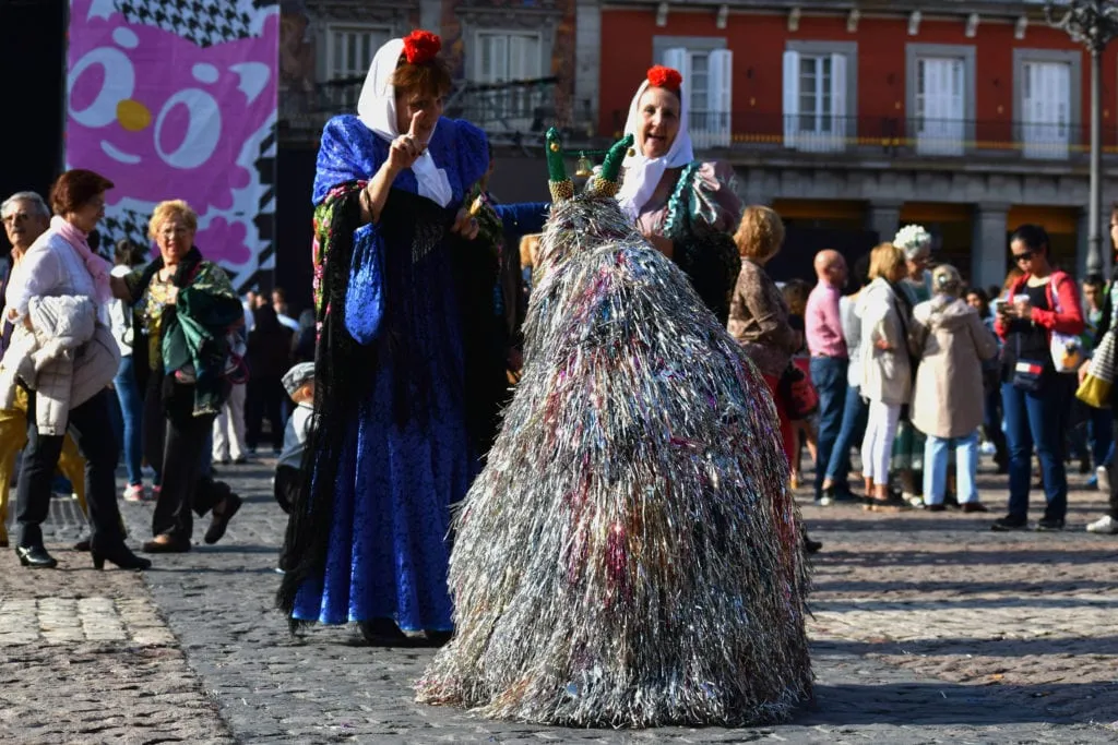 Two women in traditional dress with a dog costume made of tassles in the foreground