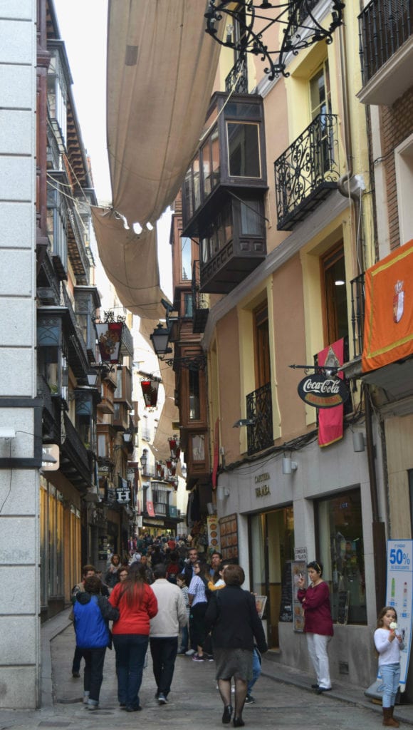 Small street in Toledo Spain with a yellow building on the right and a crowd below