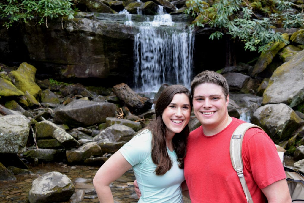 kate storm and jeremy storm standing in front of a small waterfall while hiking rainbow falls trail in gsmnp