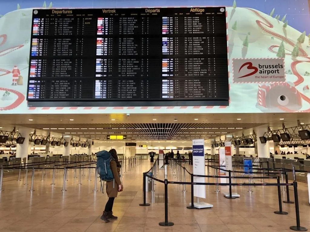 Kate Storm wearing a brown coat and blue backpack, looking up at a departures board in an airport. Her purse holds some of her long haul flight essentials!