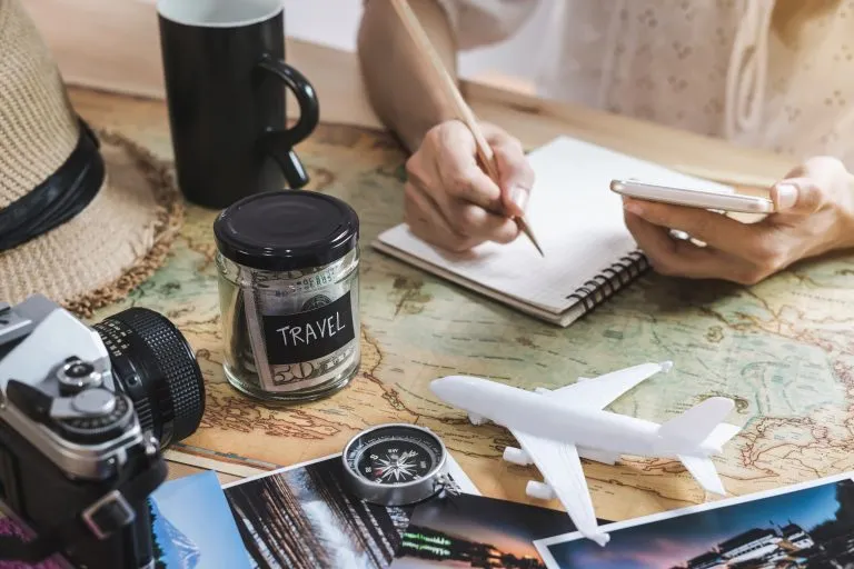 young woman travel budgeting in a notebook, with a model airplane and a jar of money in the foreground as she creates a travel budget