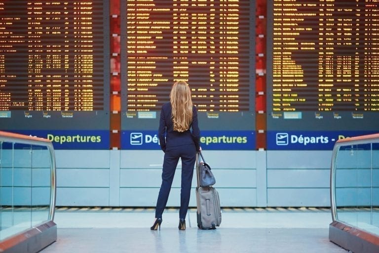 Woman with long blond hair standing in front of departures board in an airport with all travel essentials for the plane in her bag