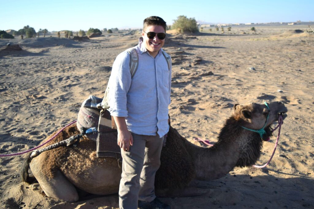 jeremy storm standing in front of a camel in morocco desert