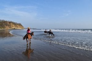 kate storm in a pink shirt horseback riding in nicaragua on a beach