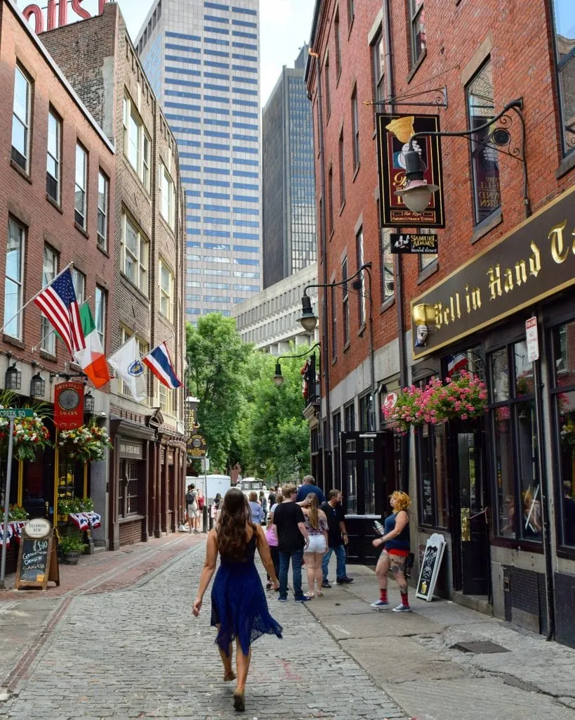 Kate Storm in a blue dress walking down a cobblestone street in Boston MA with skyscrapers ahead