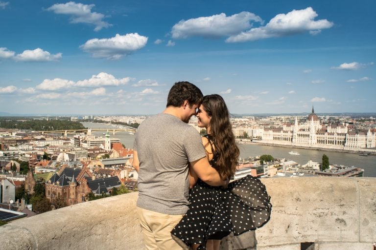 kate storm and jeremy storm posing at the top of fishermans bastion with parliament in the background in budapest hungary