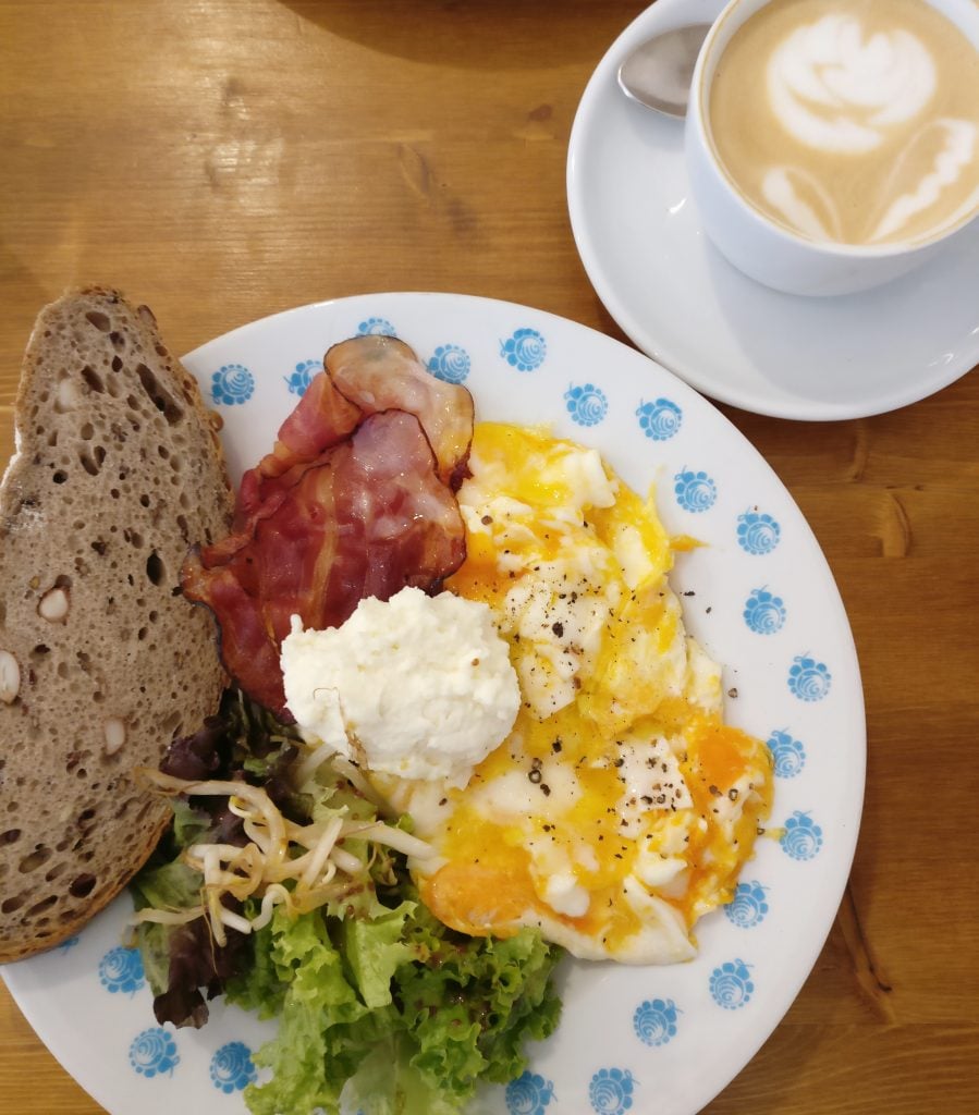 view of a scrambled egg, bacon, salad, and bread breakfast in prague with coffee shot from above