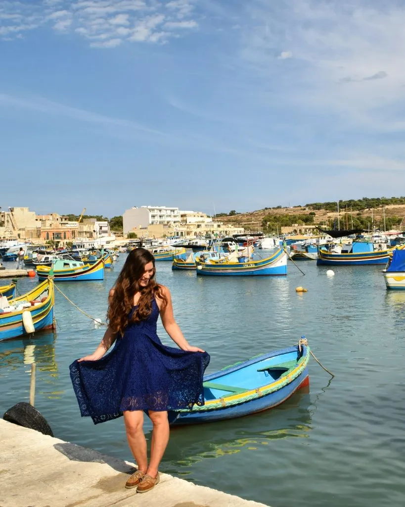 kate storm in a navy blue dress in front of the marsaxlokk harbor, one of the best places to visit in malta