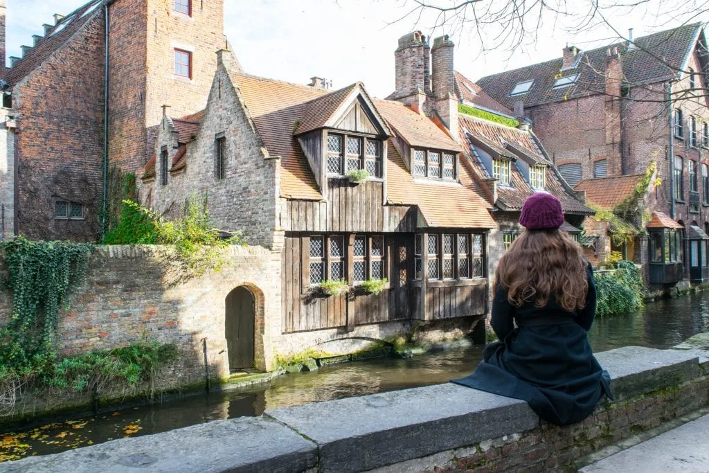 Kate sitting on a wall next to a canal in Bruges Belgium, with a wooden inn in front of her. Kate is wearing a black coat--by mid-November in Europe, you'll want a coat in many areas!