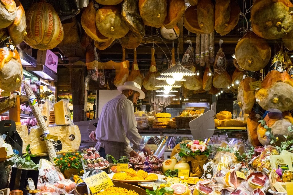 Man cutting meat at a butcher shop with his back turned to the camera. He's wearing a hat and the photo is framed by hams hanging in the shop window.