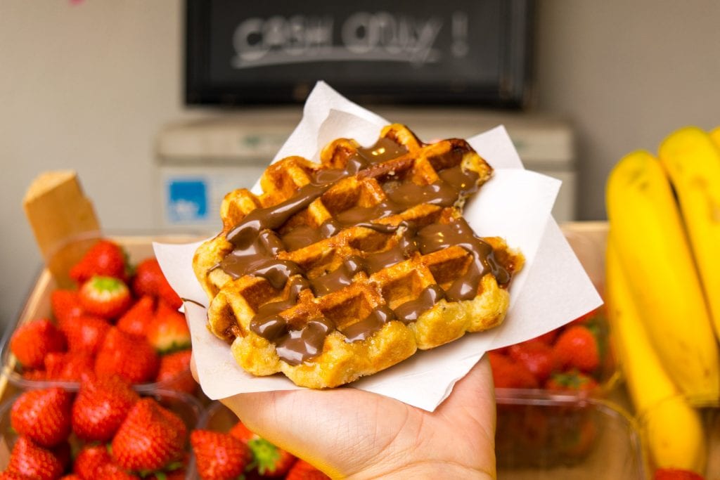 Belgian waffle being held up in front of a pile of strawberries. One bite has been taken out of the waffle.