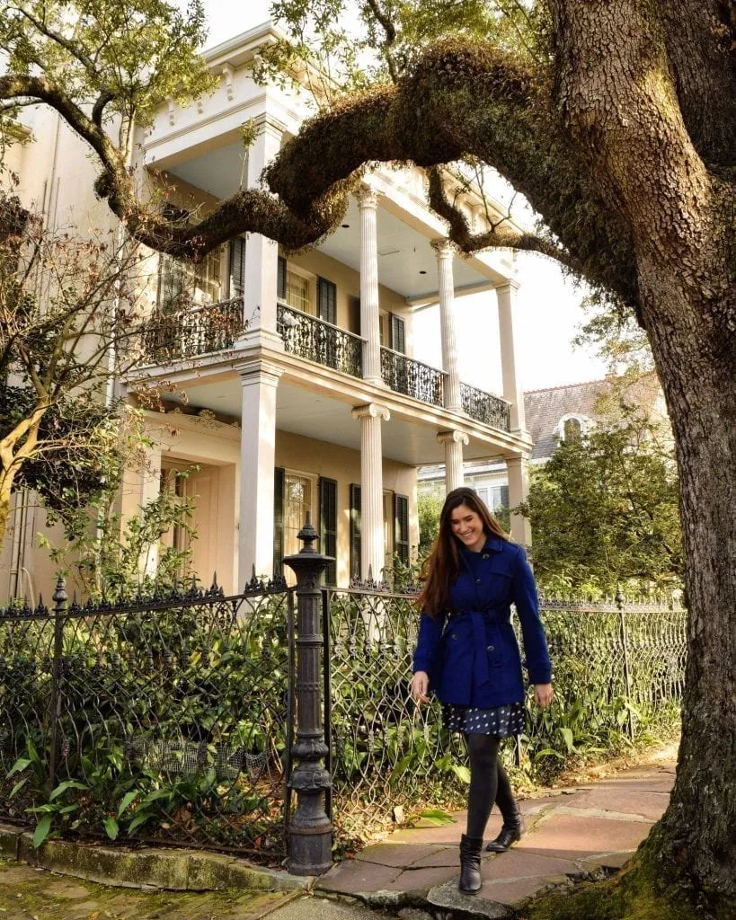 Kate Storm in a blue coat walking in front of a mansion and below a large oak tree in the Garden District during 3 days in NOLA