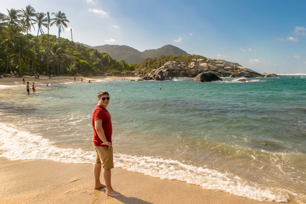 Jeremy Storm in a red shirt standing next to the water at Cabo San Juan Beach in Tayrona National Park. We definitely recommend seeing this beach as part of your 14 days in Colombia itinerary!